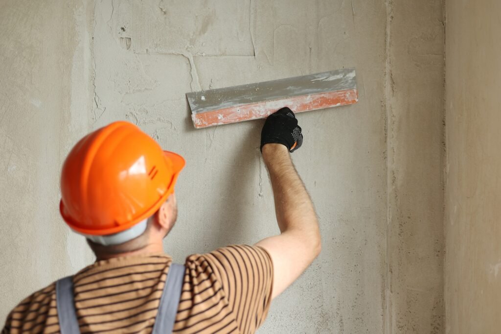 Rear view of handyman worker in overalls and protective helmet plastering concrete wall with putty u